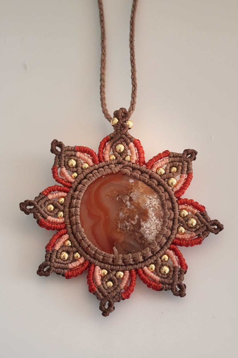 A handmade macrame pendant necklace featuring a central round Carnelian Agate cabochon surrounded by a sunflower mandala medallion design of layered rust, peach, and brown woven cords with small brass beads, hanging against a plain light background.