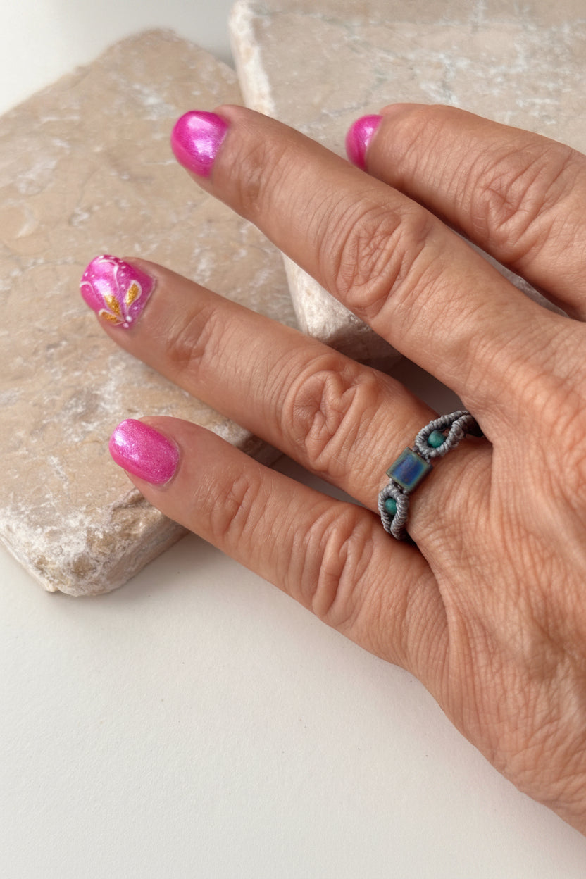 A handmade minimalist gray macrame ring with an iridescent blue-purple square Miyuki Tila bead at the center and two teal seed bead accents, worn on a finger resting on a pink stone surface with pink nail art in the background