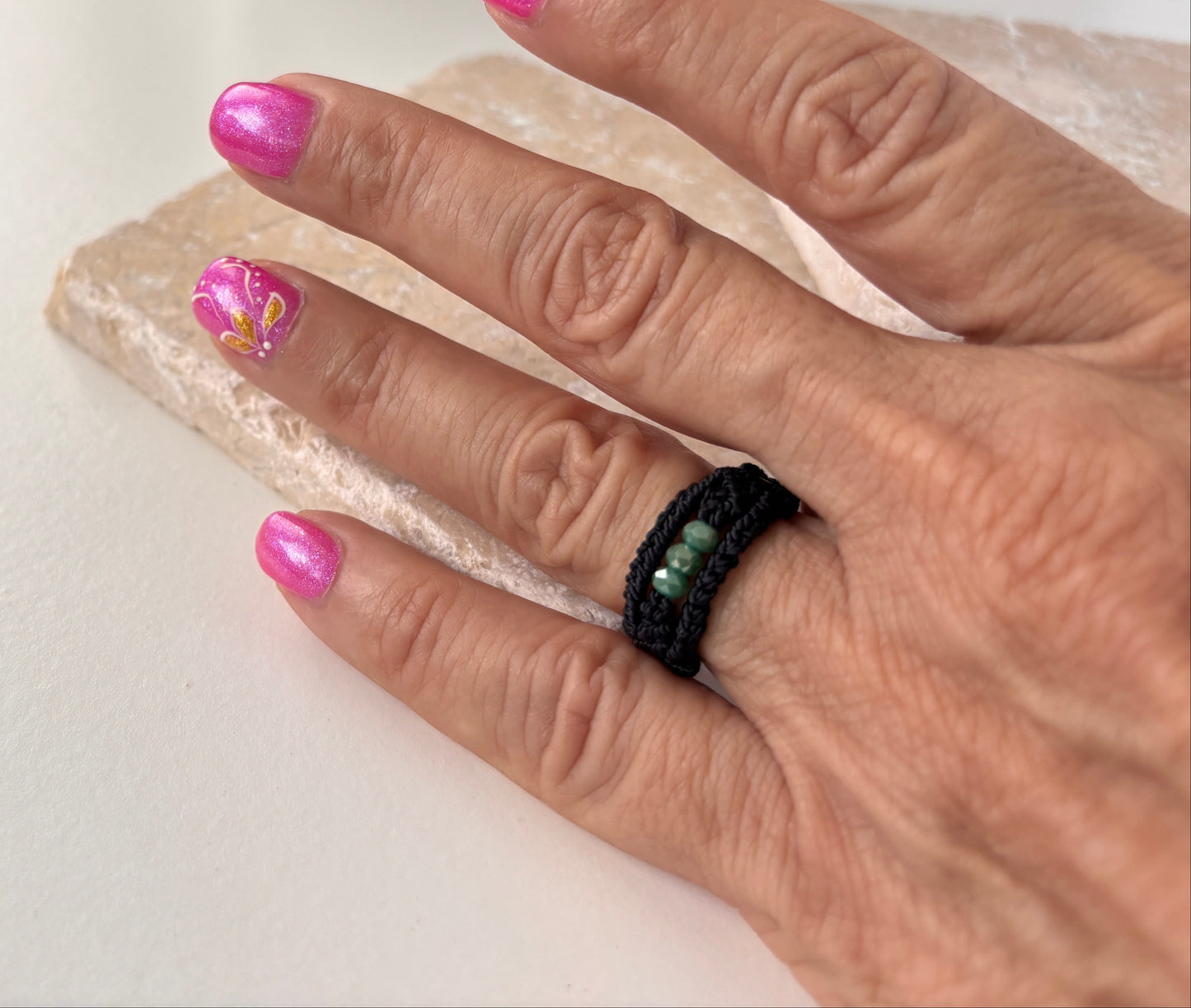 A handmade macrame band ring in black flat waxed nylon cord with a double-wrap braided design and three faceted mint green glass crystal beads, worn on a finger resting on a white stone surface with pink nail art in the background.