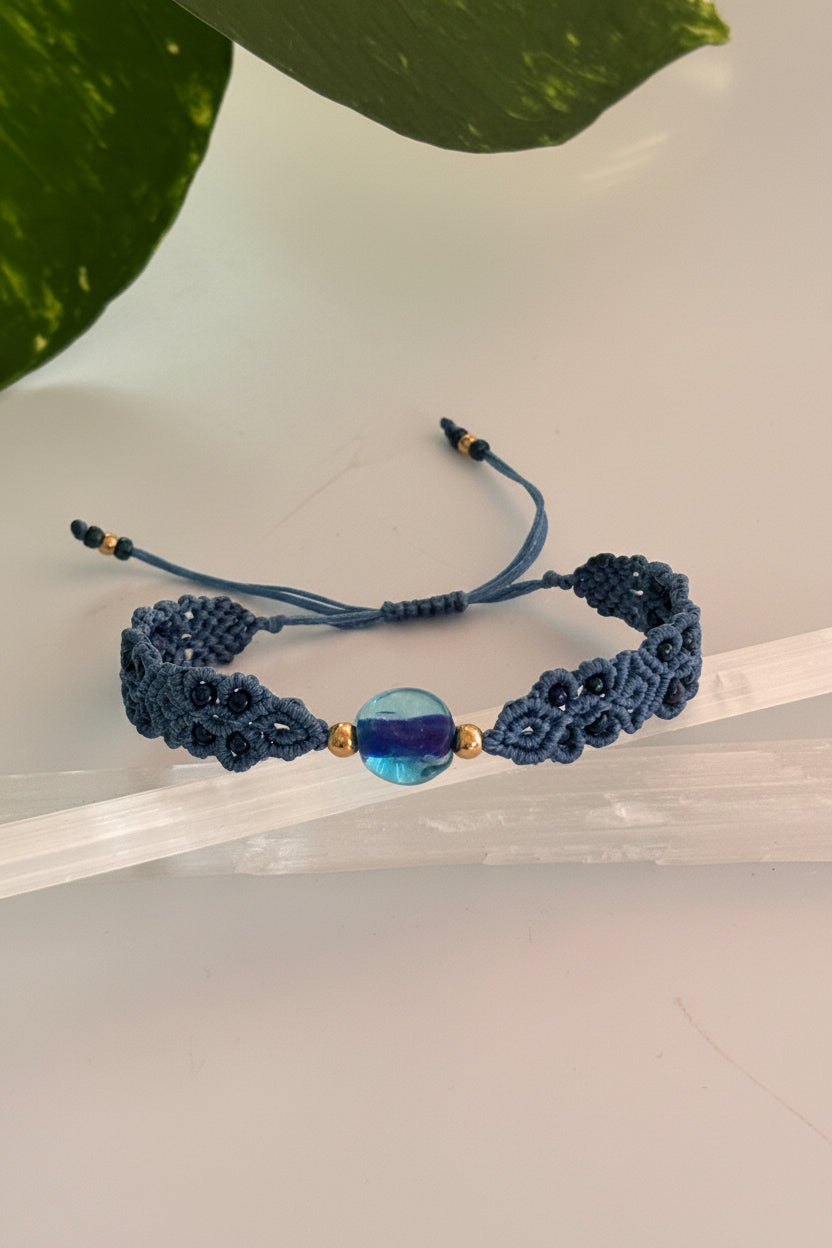 A handmade macrame bracelet in a medium blue with a central aqua blue glass disc bead and small brass spacer beads, displayed on a clear acrylic block.