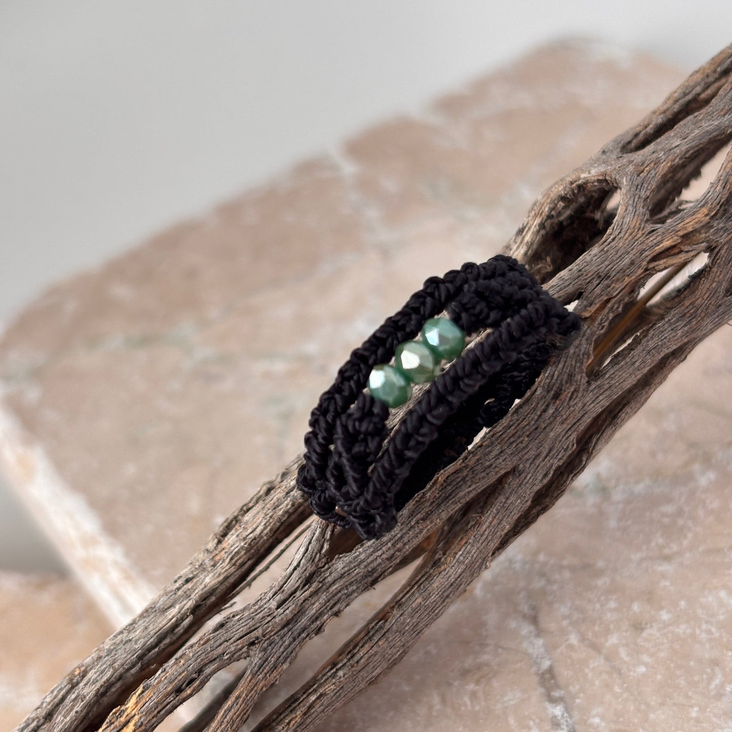 A close-up of a handmade macrame band ring in black flat waxed nylon cord with a double-wrap braided design and three faceted mint green glass crystal beads across the center face, resting on weathered driftwood against a pink stone surface.
