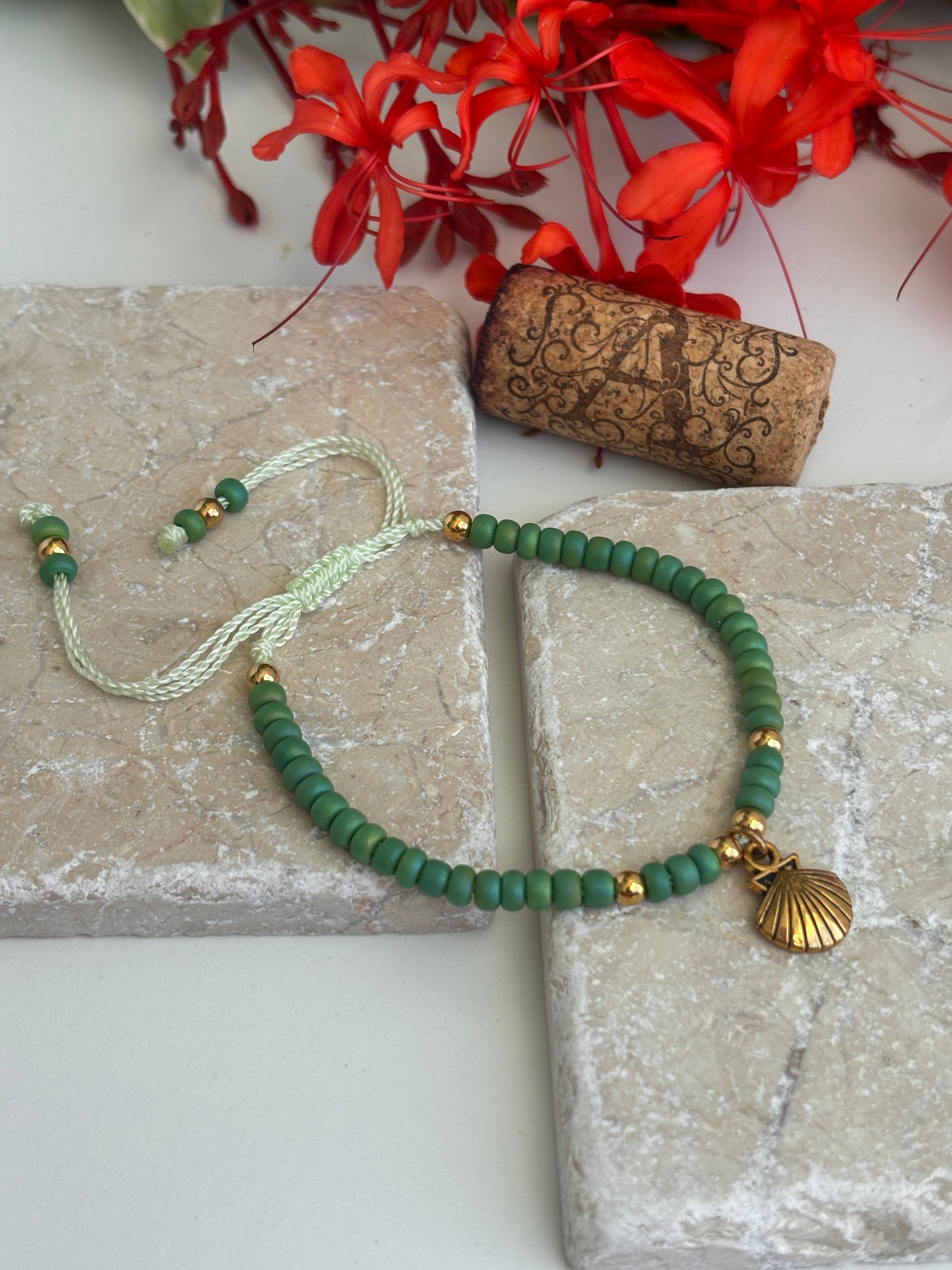 A green beaded bracelet with a  brass shell charm and brass spacer beads, displayed on a textured stone block next to a cork stopper and red flowers.