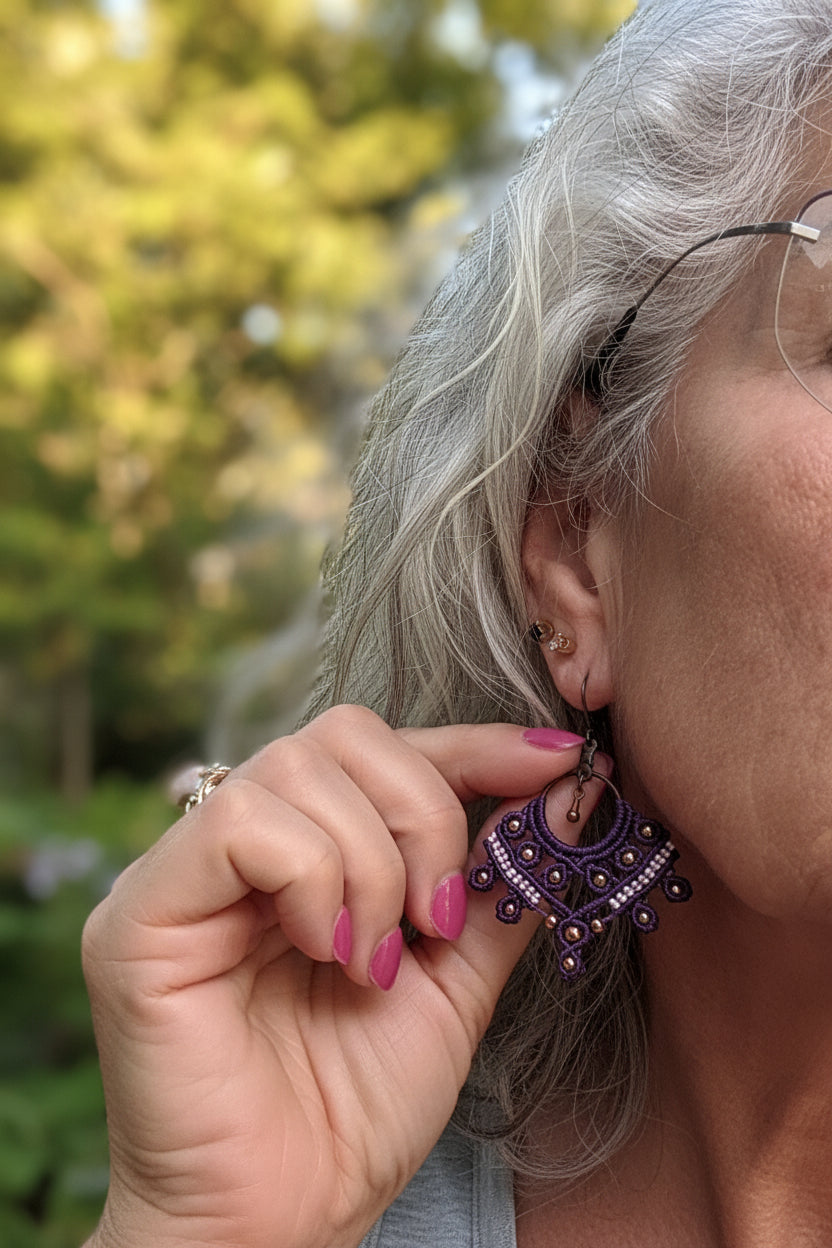A woman with silver hair wearing a handcrafted macrame chandelier earring in deep purple nylon cord with copper round beads, a lavender macrame cord accent row, and an antique copper lever back ear wire, shown in an outdoor garden setting with soft-focus green and yellow foliage in the background