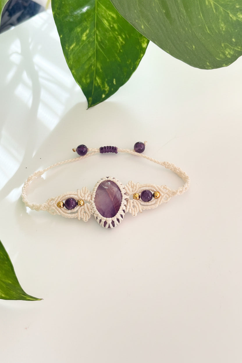 A handmade macrame bracelet with a central oval purple stone, small purple beads, and tiny gold accent beads, displayed on a white surface with green leaves in the background.
