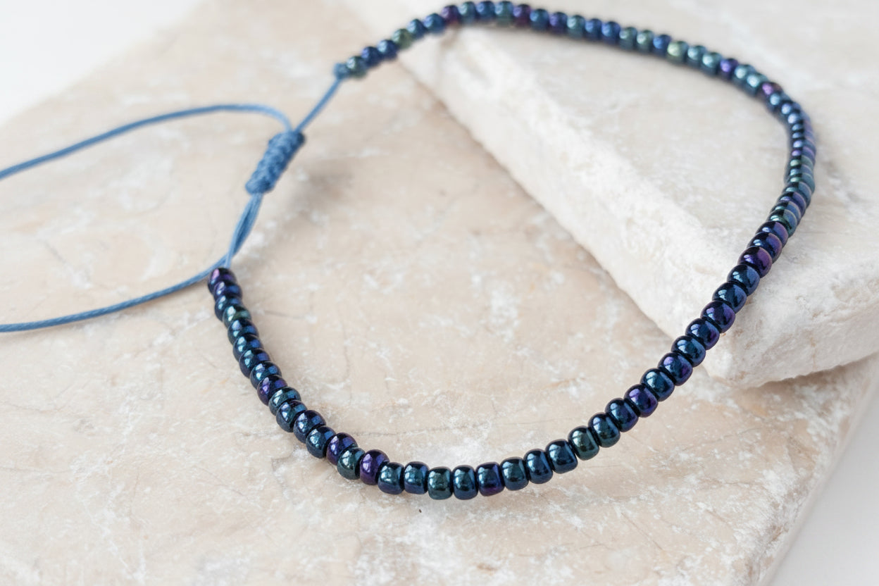 A close-up of a handcrafted single strand bracelet with size 8/0 dark blue iridescent seed beads showing a flash of purple and blue shimmer, on a medium blue nylon cord with an adjustable slide closure, resting on a white selenite crystal slab