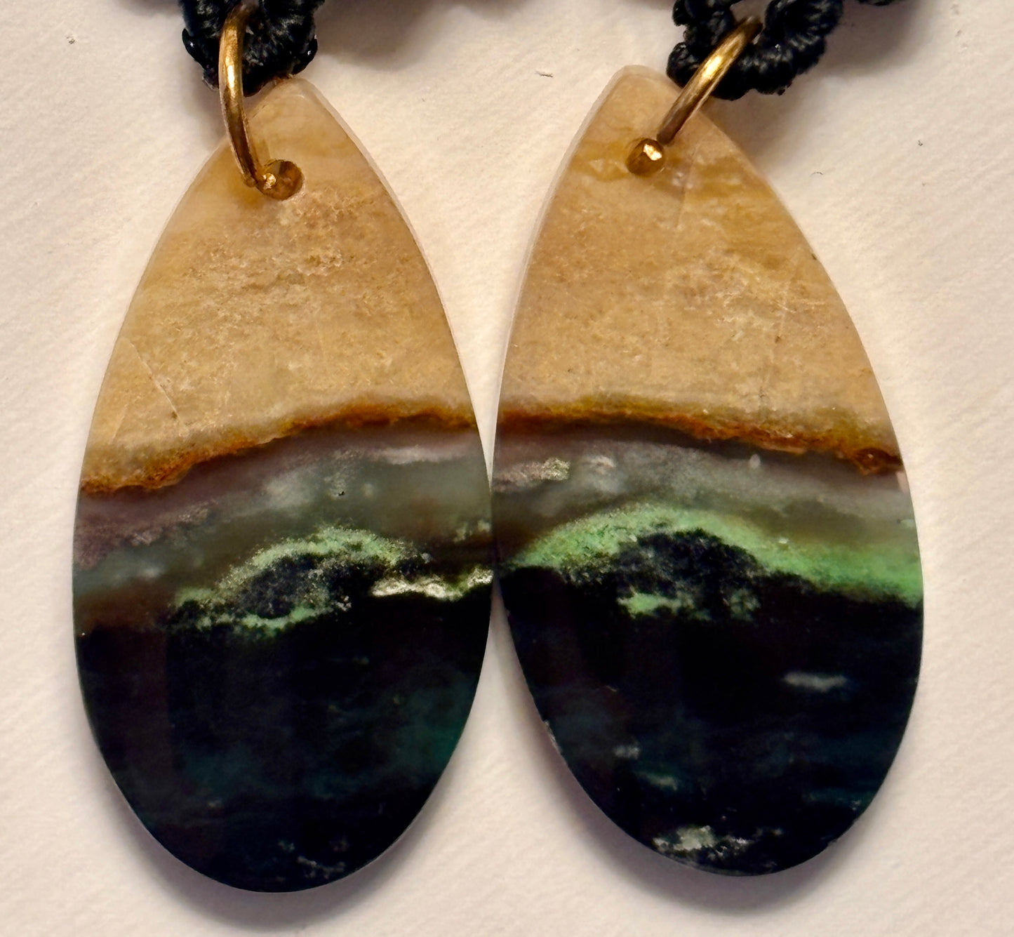 Macro close-up of handmade black macrame drop earrings showing green chalcedony teardrop gemstone detail with natural tan, green, and black mineral banding, and gold-tone jump ring connector.
