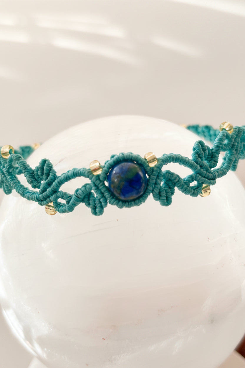 A teal macrame bracelet with a central dark blue stone and small gold-lined seed beads, displayed on a white crystal sphere.