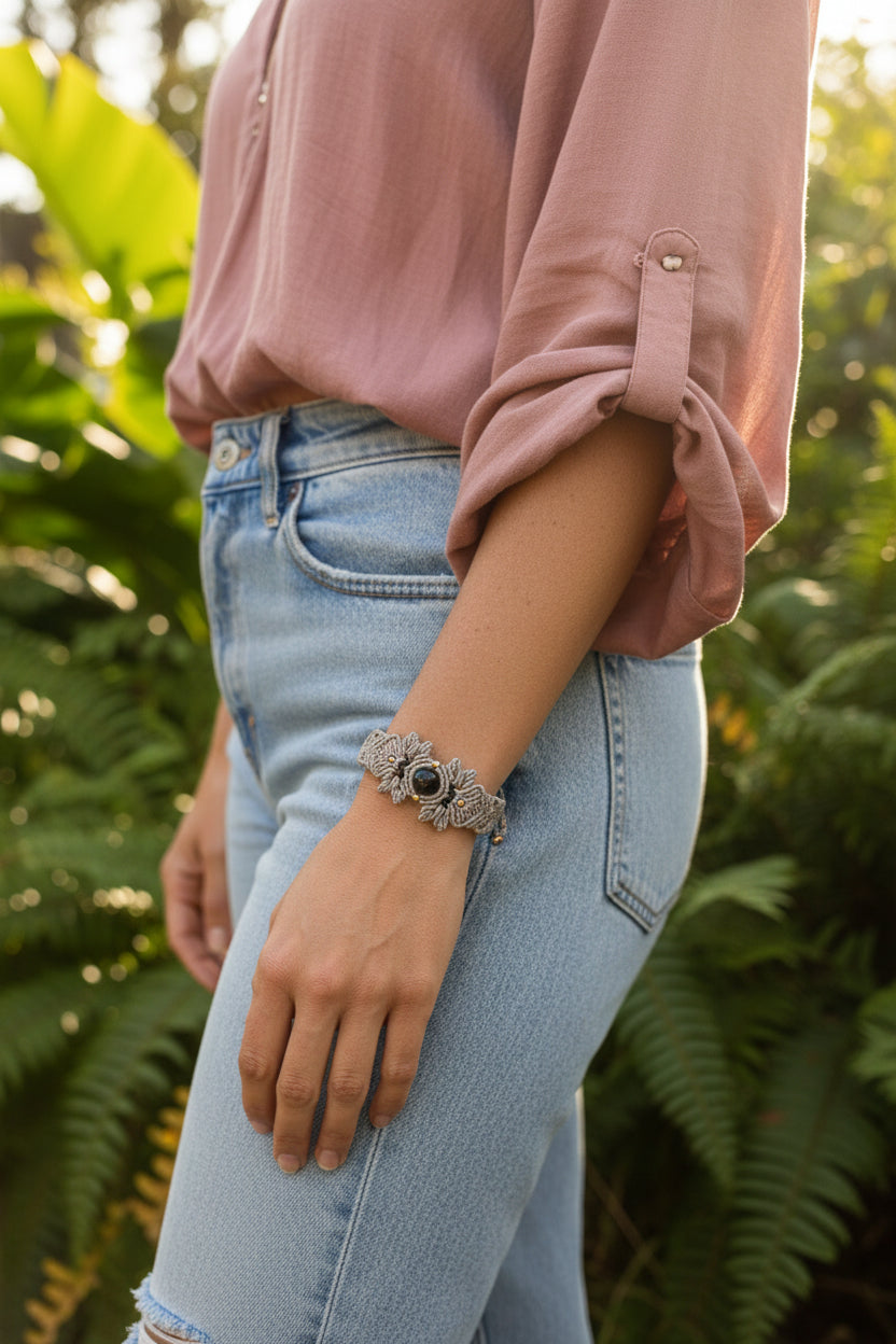 A handmade gray macrame bracelet with a central lapis lazuli stone and small brass beads, displayed on a woman's wrist,  She is dressed in blue jeans and a rose colored blouse while standing outdoors near greenary.