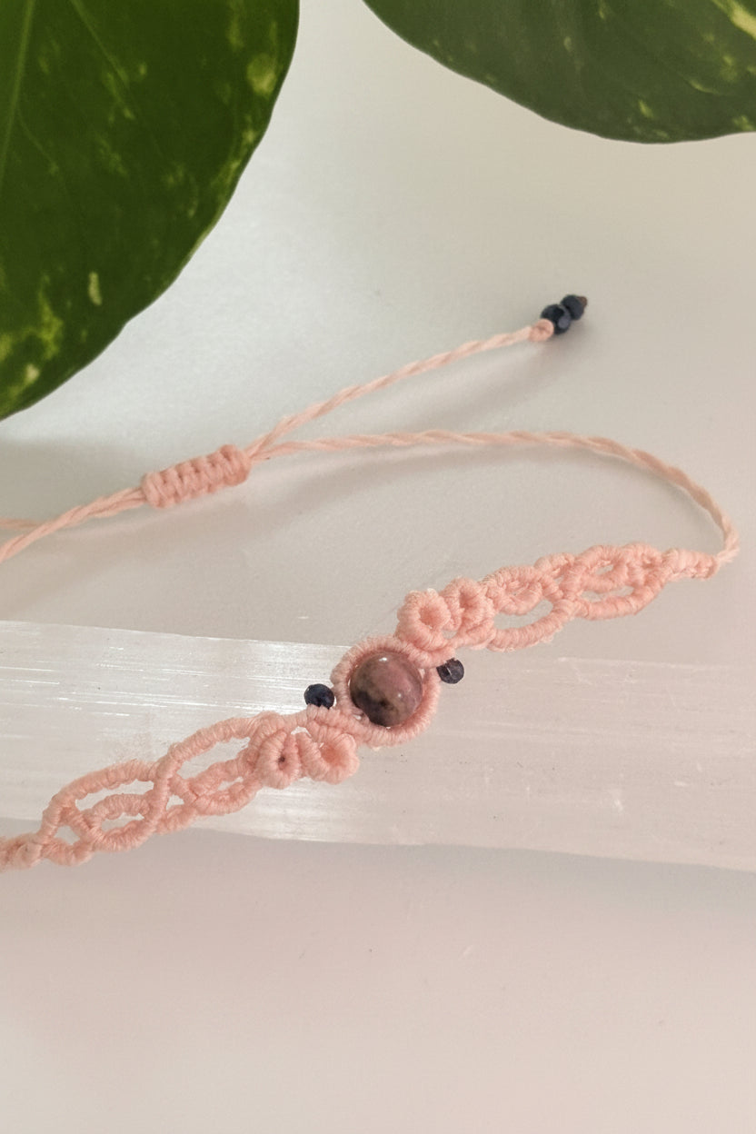 A light pink macrame bracelet with a central Rhodonite stone bead and black iridescent crystal beads, displayed on a clear acrylic block.