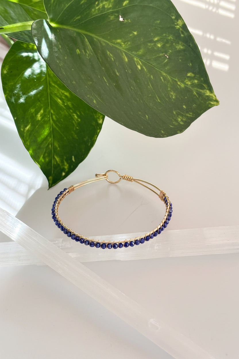 A brass bangle bracelet with a row of small blue faceted crystal beads, hanging on a clear plastic hanger next to a green houseplant leaf.