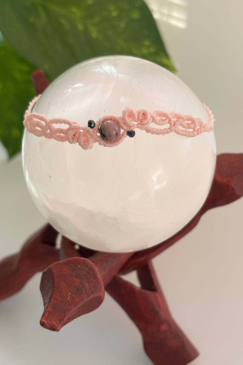 A light pink macrame bracelet with a central Rhodonite bead and two black iridescent crystal beads, displayed on a clear crystal sphere resting on a dark reddish-brown wooden stand.