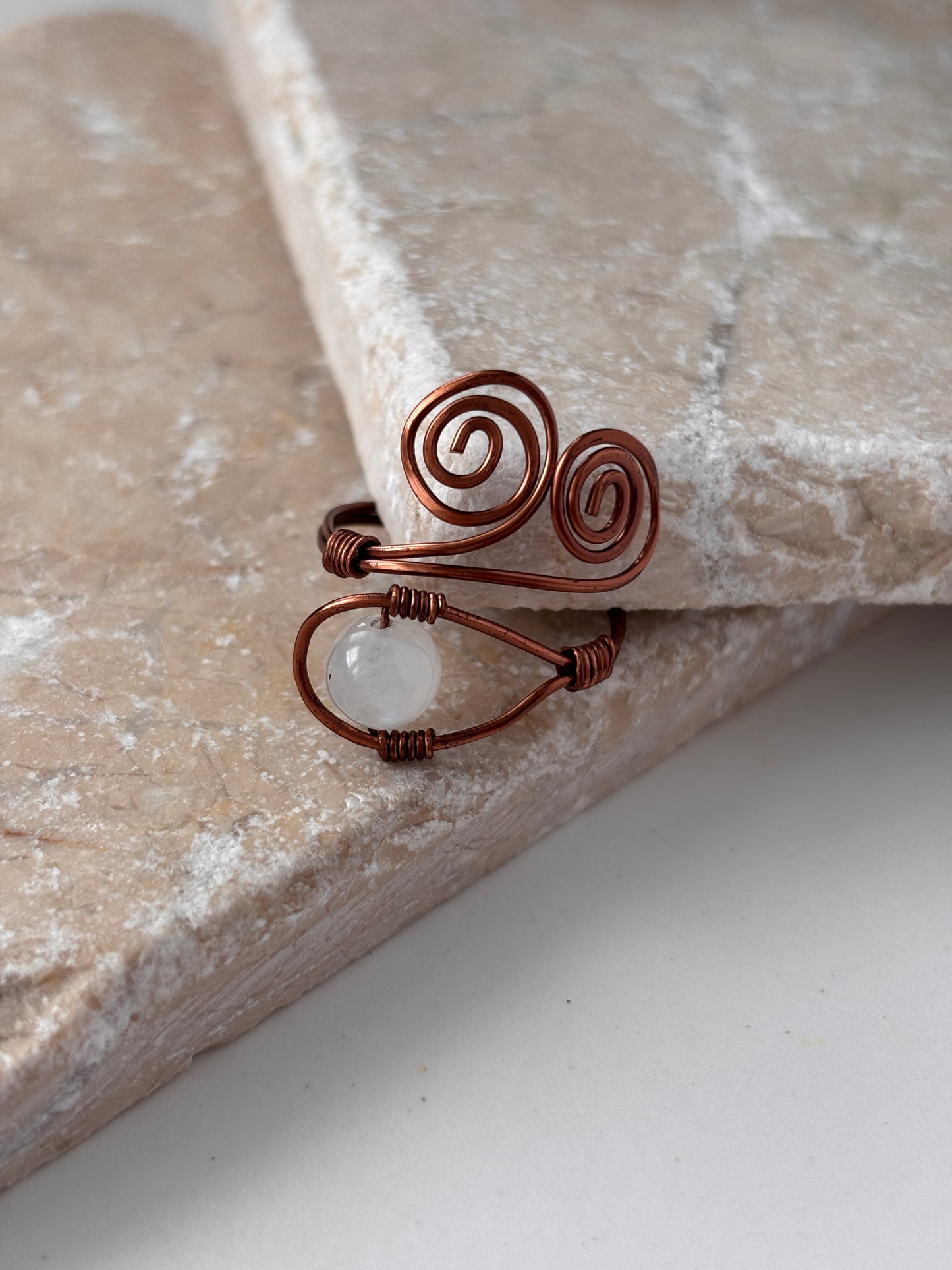 A handcrafted copper wire ring with a double spiral flourish and a wire-wrapped round moonstone bead at the base, leaning against the edge of a natural stone slab against a white background.