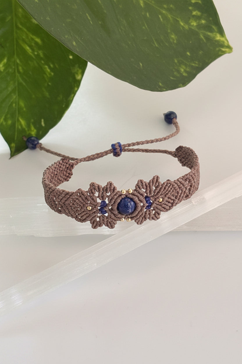 A brown macrame bracelet with a central floral design featuring lapis lazuli stones and small brass beads, displayed on a clear crystal block with green leaves in the background.