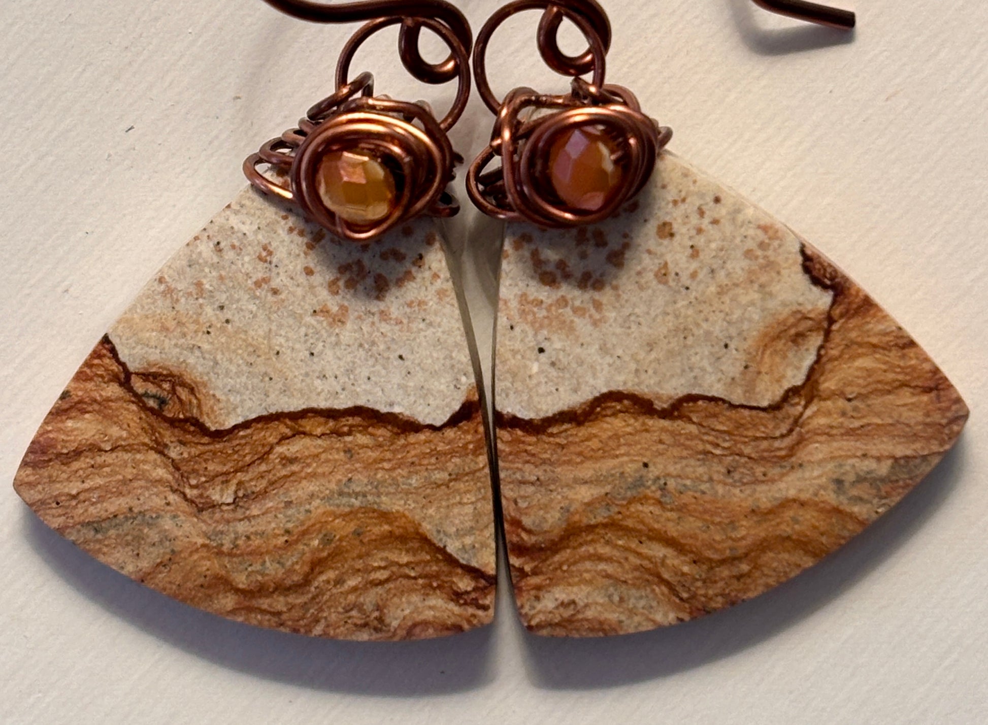 Macro close-up of handmade petrified wood fan-shaped drop earrings showing natural cream and caramel wood grain banding, iridescent orange faceted crystal bead, and intricate antique copper wire wrap bail detail.