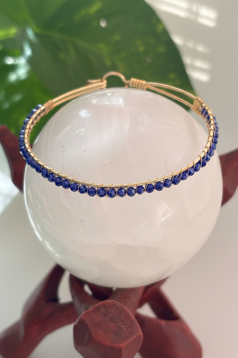 A brass bangle bracelet decorated with a single row of blue faceted crystal beads, displayed on a white crystal sphere stand.