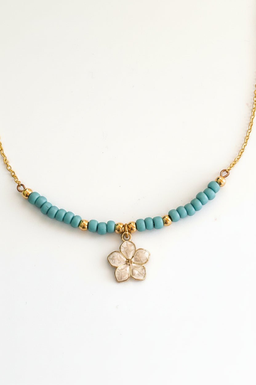A delicate brass chain necklace featuring a row of small turquoise blue seed beads with brass spacer accents and a central brass white enamel flower charm, displayed on a neutral light gray background.