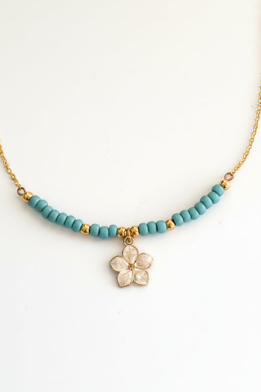 Turquoise seed bead necklace with brass chain, brass spacer beads, and an ivory enamel flower charm on a light gray background.