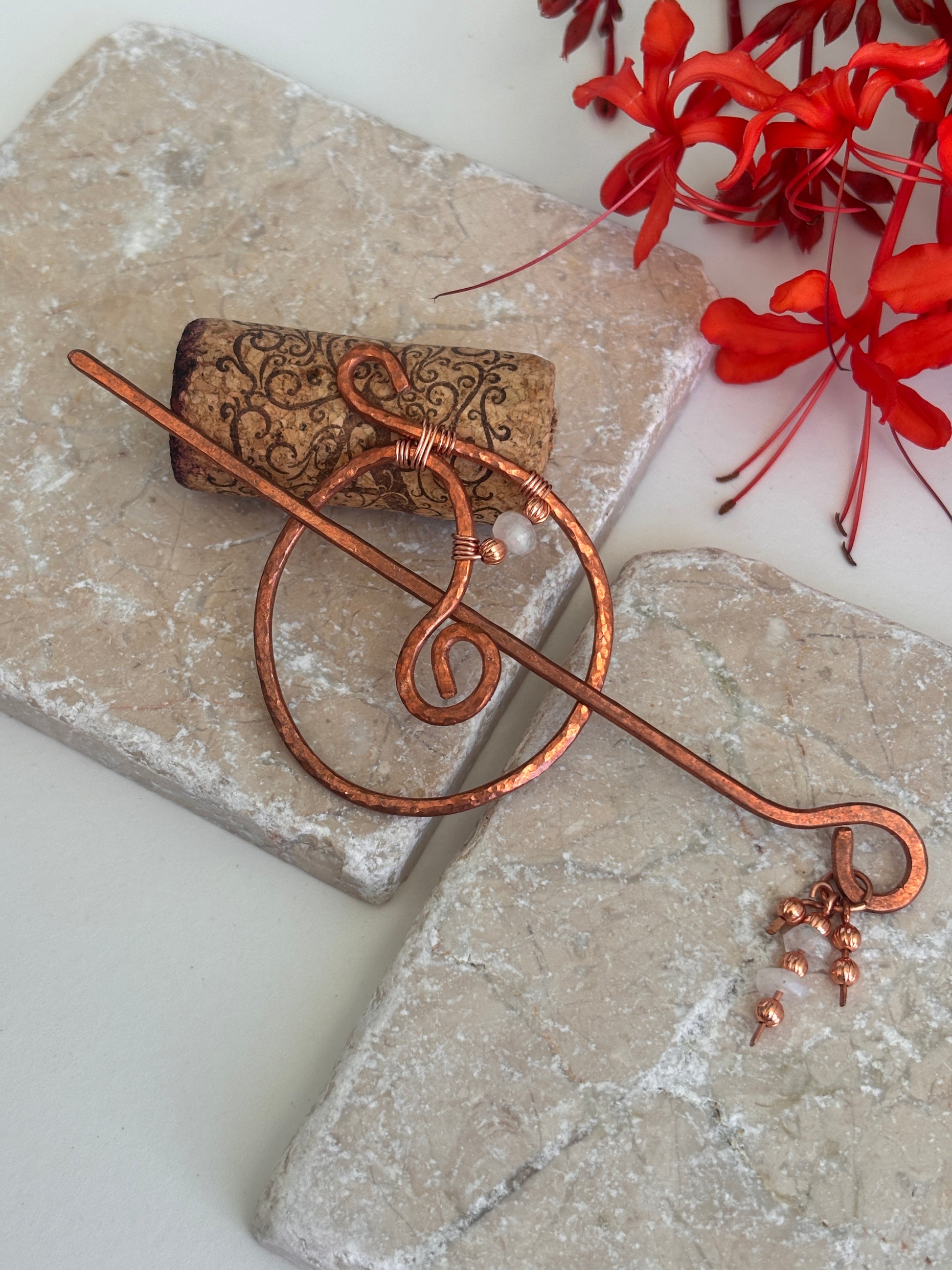 A hammered copper hair stick styled with a decorative wire design and small dangling copper and moonstone beads, resting on textured stone tiles with red flowers in the background.
