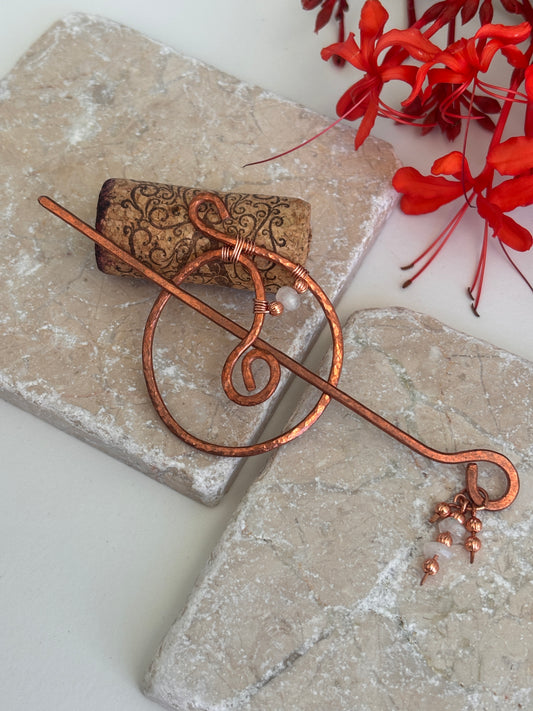 A hammered copper hair stick styled with a decorative wire design and small dangling copper and moonstone beads, resting on textured stone tiles with red flowers in the background.