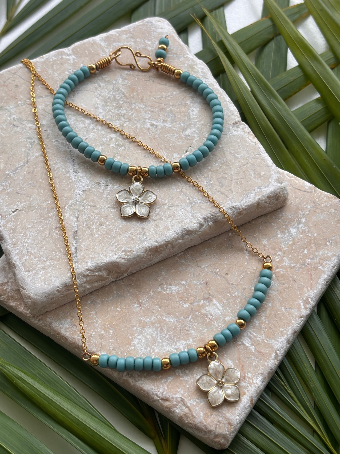 A jewelry set featuring a turquoise blue and brass beaded necklace with a white enamel flower charm, a matching beaded bracelet, styled on stacked stone slabs with green palm leaves.  Each item sold separately.