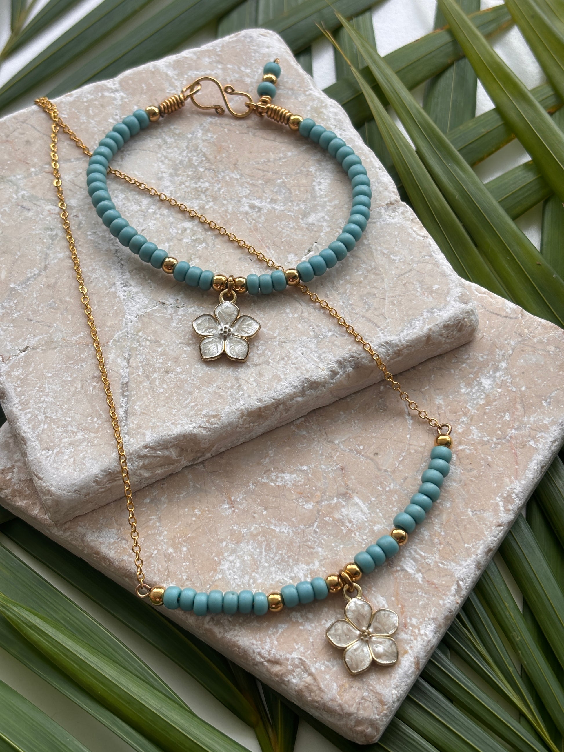A jewelry set featuring a turquoise blue and brass beaded necklace with a white enamel flower charm, a matching beaded bracelet, styled on stacked stone slabs with green palm leaves.  Each item sold separately.