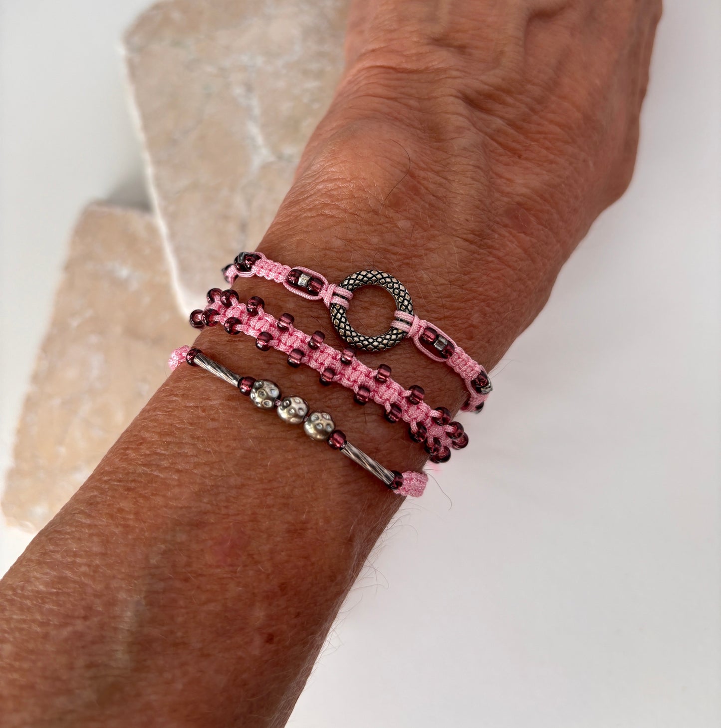 A side view of three pink macrame bracelets stacked on a wrist against a white stone background — one with a silver-tone open ring centerpiece, one with cranberry seed beads woven along the edges, and one with a silver-tone tube bead and silver-tone ball accent beads on pink nylon cord.