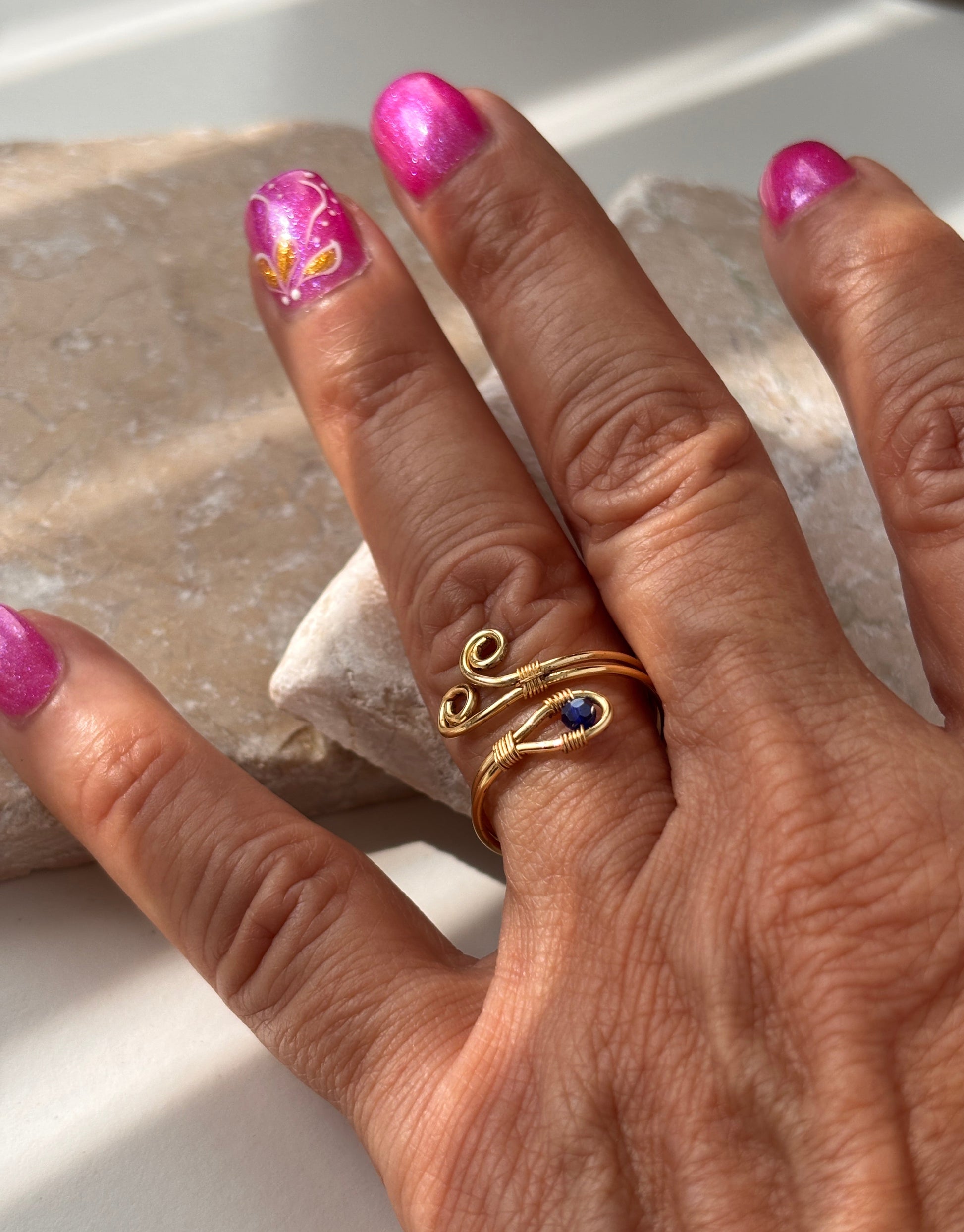 A handcrafted gold-tone wire ring with a double spiral flourish and a 3mm deep blue faceted crystal bead, worn on a finger resting on a natural stone slab in natural sunlight, with pink nail art in the background