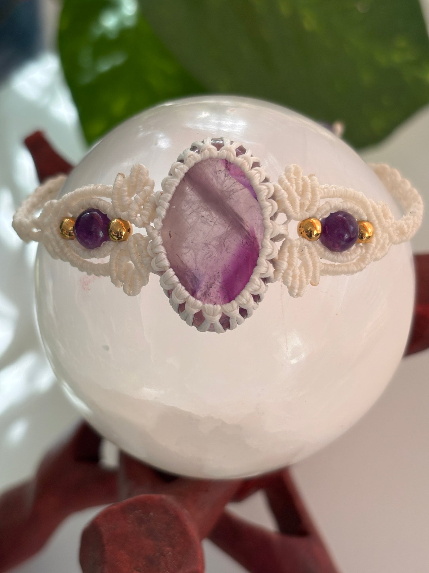 A handmade macrame bracelet with a central oval purple amethyst stone and two smaller round purple amethyst beads, displayed on a white crystal sphere.