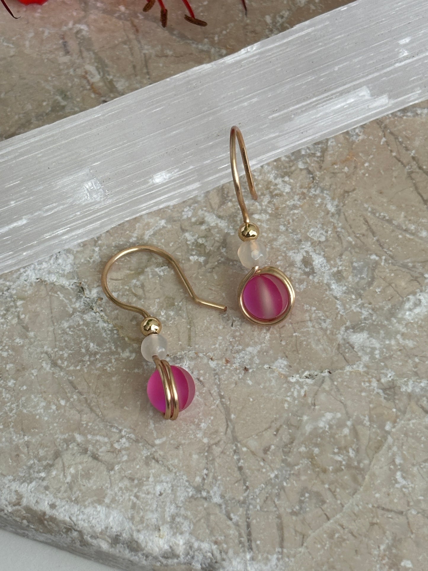 A pair of brass dangle earrings with vivid fuchsia round glass beads and small faceted white agate beads, displayed on a textured beige stone surface with a translucent white crystal slab behind them.