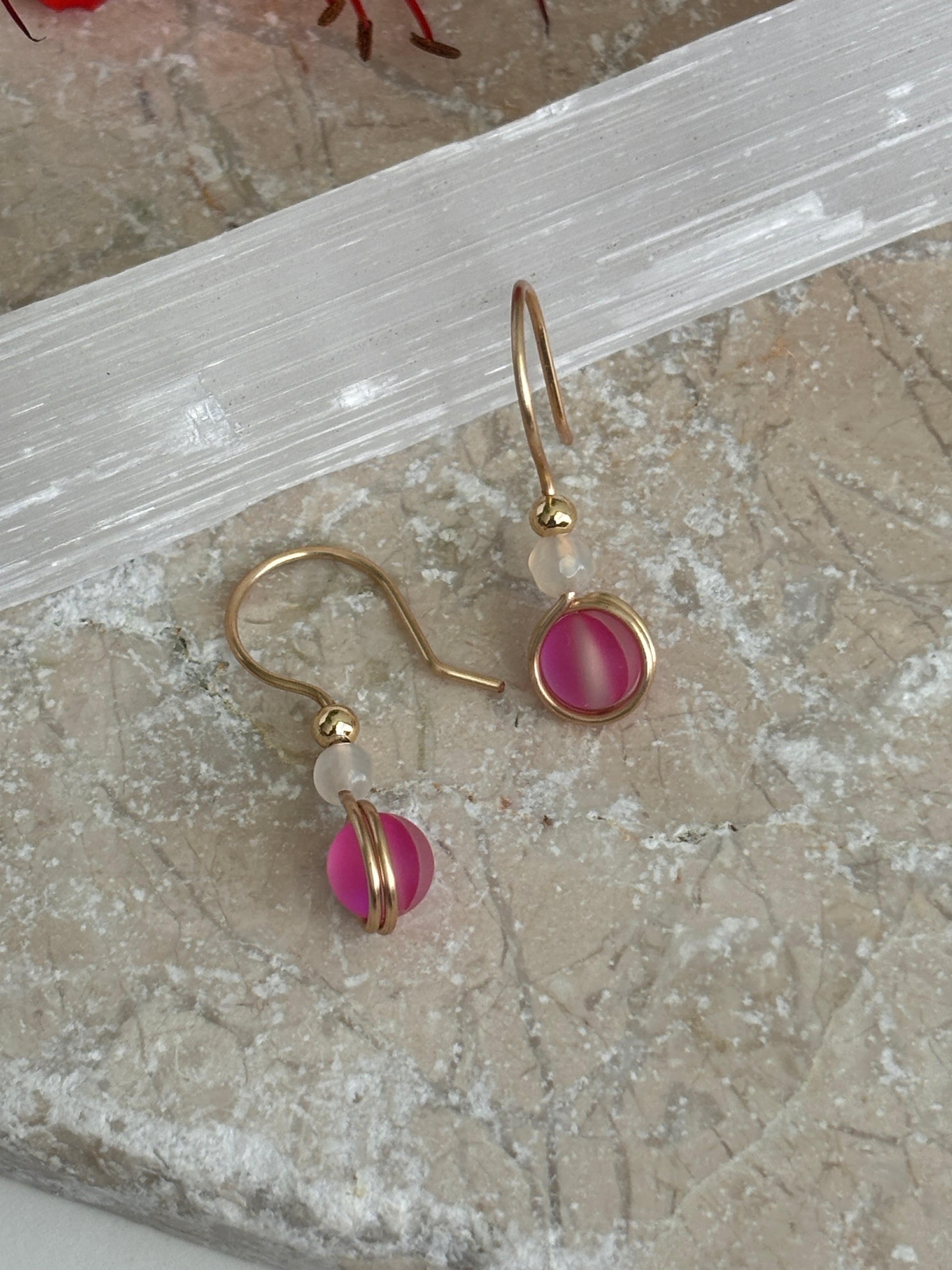 A pair of brass dangle earrings with vivid fuchsia round glass beads and small faceted white agate beads, displayed on a textured beige stone surface with a translucent white crystal slab behind them.