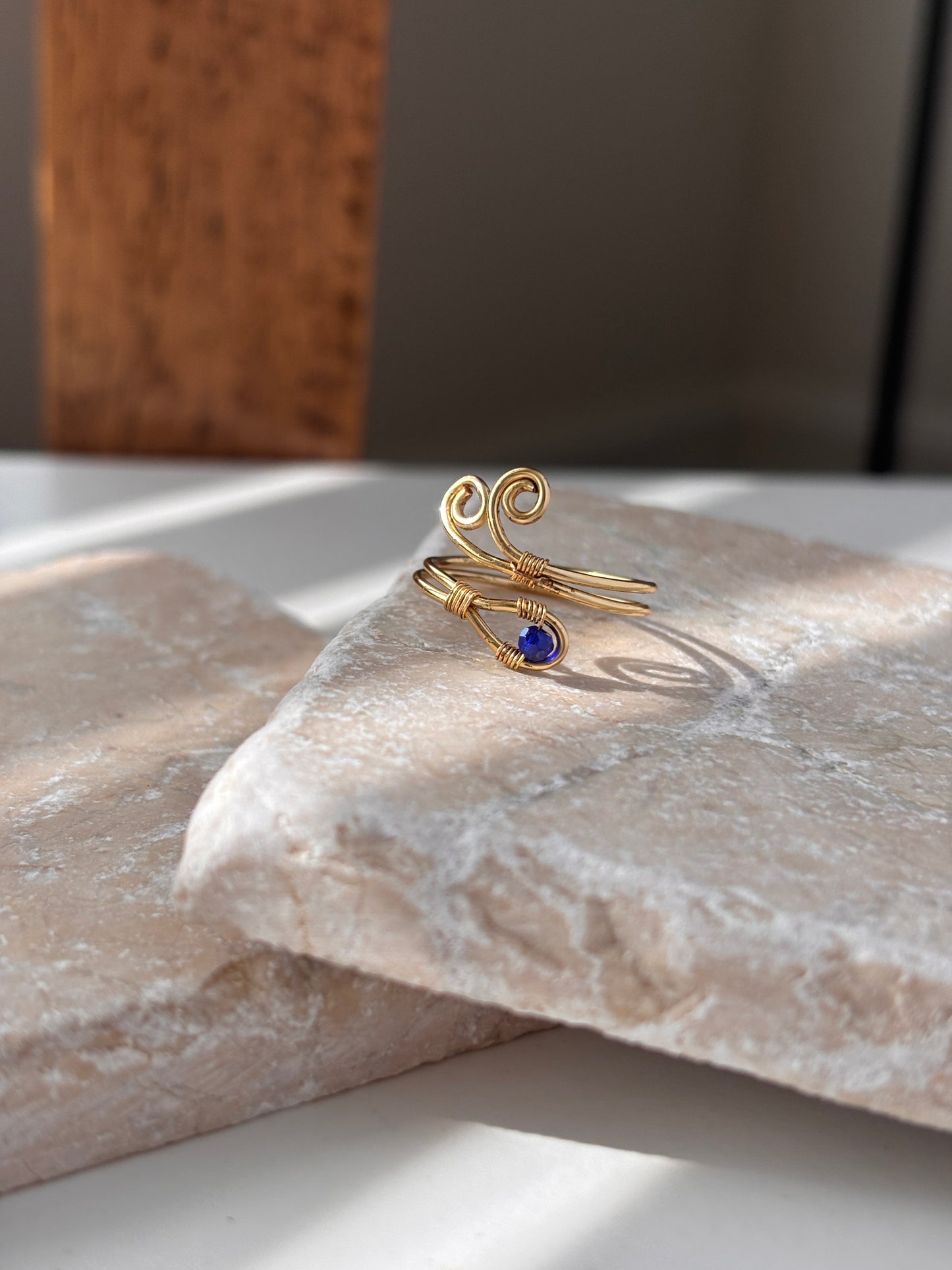 A handcrafted gold-tone wire ring with a double spiral flourish and a 3mm deep blue faceted crystal bead wire-wrapped at the base, resting on a natural stone slab in soft natural sunlight showing the open adjustable band tail, against a white background with driftwood in the soft-focus background