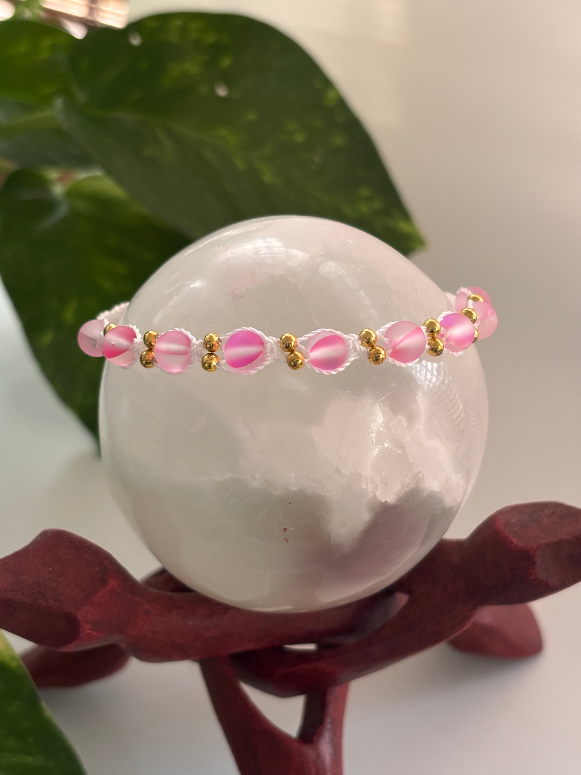 A delicate beaded macrame bracelet with frosted pink round glass beads and small brass spacer beads, displayed on a white crystal sphere atop a dark red wooden stand, with green leaves in the background.