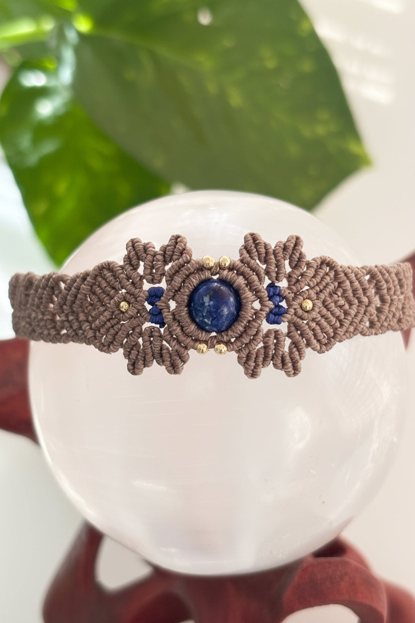 A handmade macrame bracelet in brown with a central lapis lazuli stone and small brass beads, displayed on a white crystal sphere.