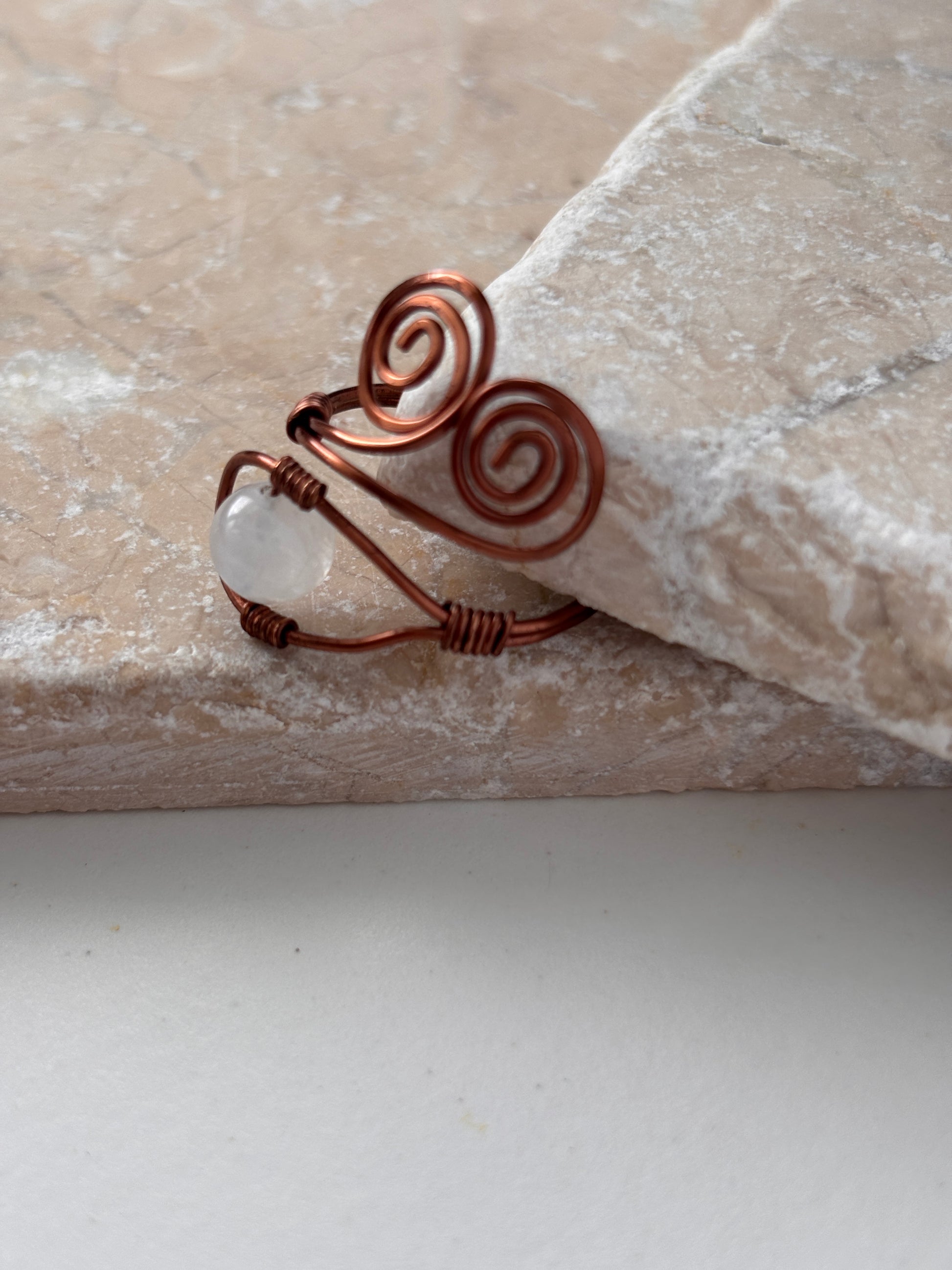A side view of a handcrafted copper wire ring with a double spiral flourish and a wire-wrapped round moonstone bead at the base, leaning against the edge of a natural stone slab against a white background.