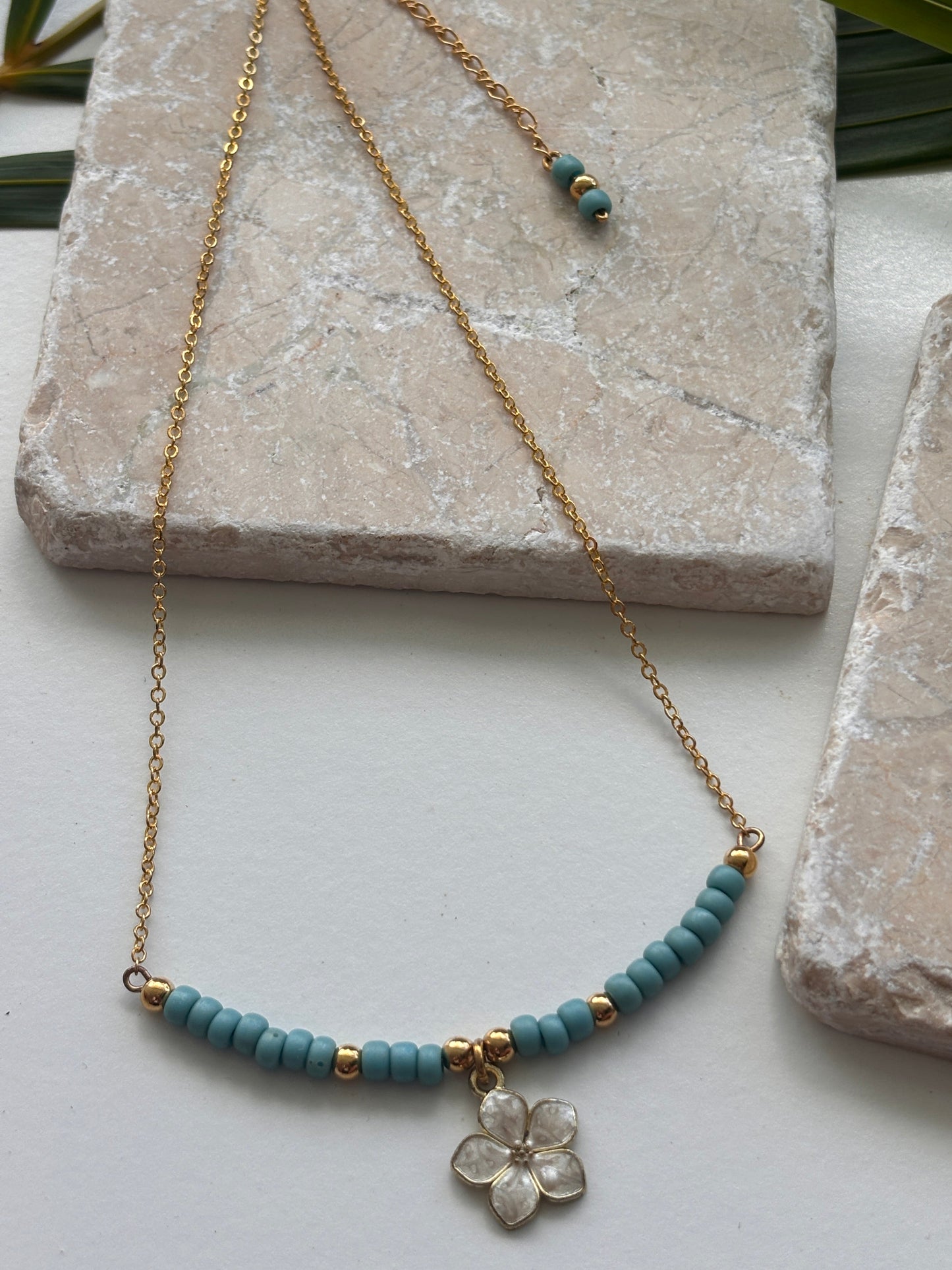 A brass chain necklace with turquoise blue beads, small brass spacer beads, and a white enamel flower pendant, styled on a white surface with stone blocks and green leaves.