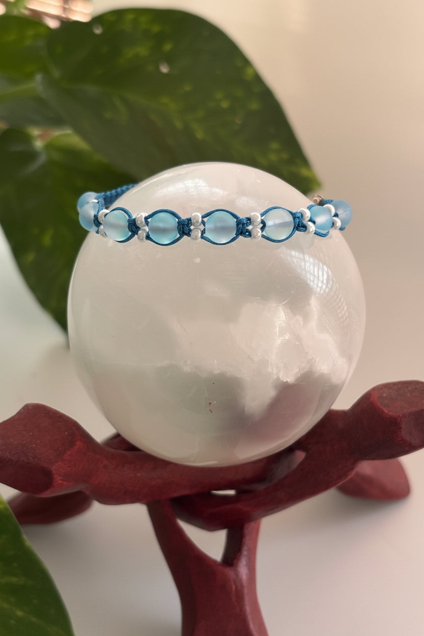 An aqua beaded macrame bracelet with teal cord and white pearl seed bead accents, displayed on a large white crystal sphere resting on a dark red wooden stand, with green leaves in the background.