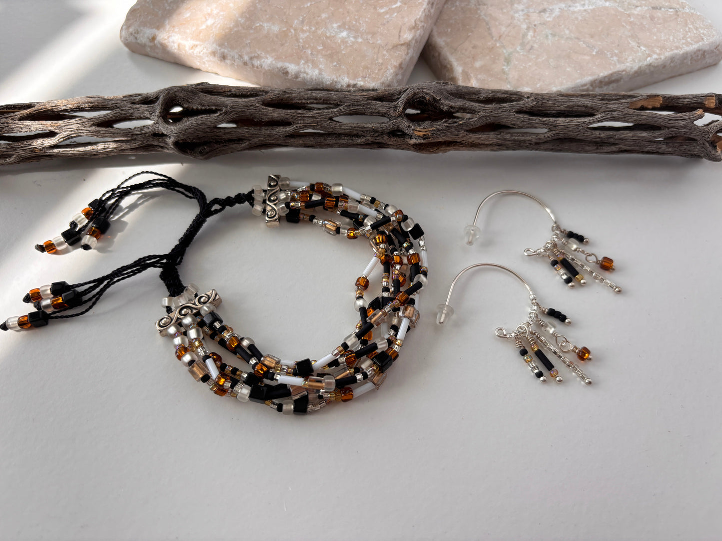 A pair of handcrafted silver-tone hoop earrings with five wire-wrapped dangle strands of black bugle beads, clear seed beads, frosted white disc beads, and an amber faceted cube bead drop, displayed alongside a seven strand seed bead bracelet in black nylon macrame cord on a white surface with pink stone slabs and driftwood. Items sold separately.