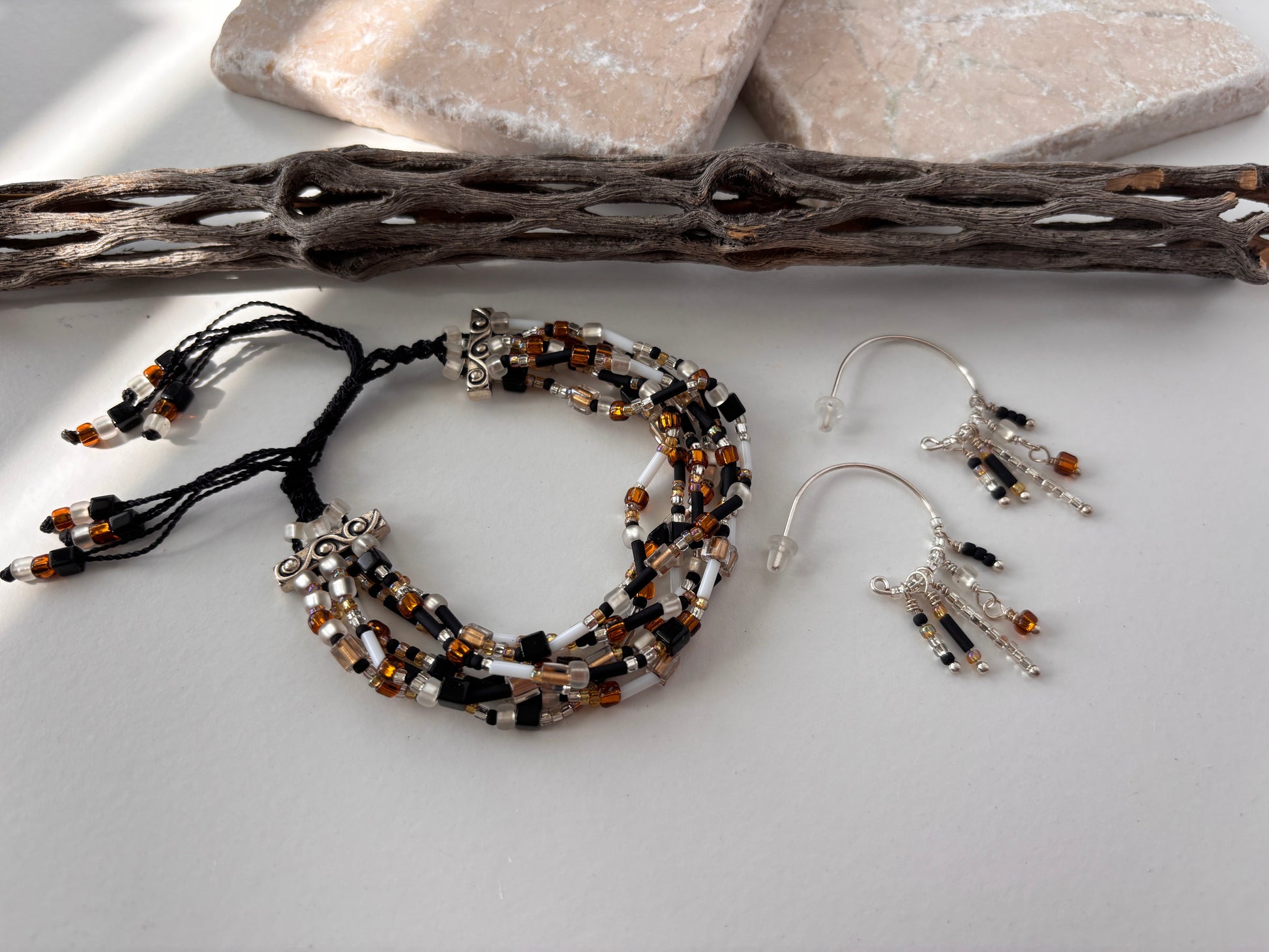 A pair of handcrafted silver-tone hoop earrings with five wire-wrapped dangle strands of black bugle beads, clear seed beads, frosted white disc beads, and an amber faceted cube bead drop, displayed alongside a seven strand seed bead bracelet in black nylon macrame cord on a white surface with pink stone slabs and driftwood. Items sold separately.
