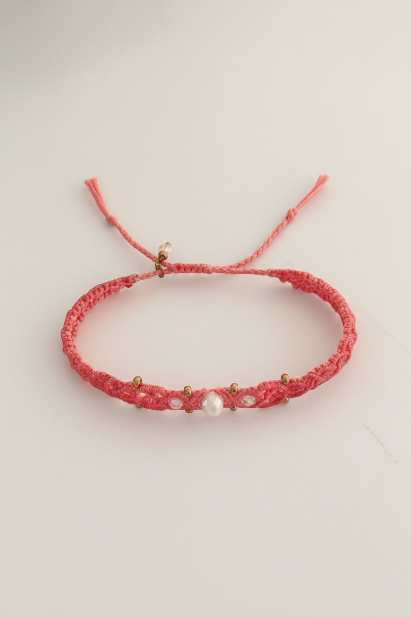 A salmon pink macrame bracelet with a iridescent faceted crystal bead, small brass spacer beads, and small iridescent pink crystal accents, displayed on a light beige background.