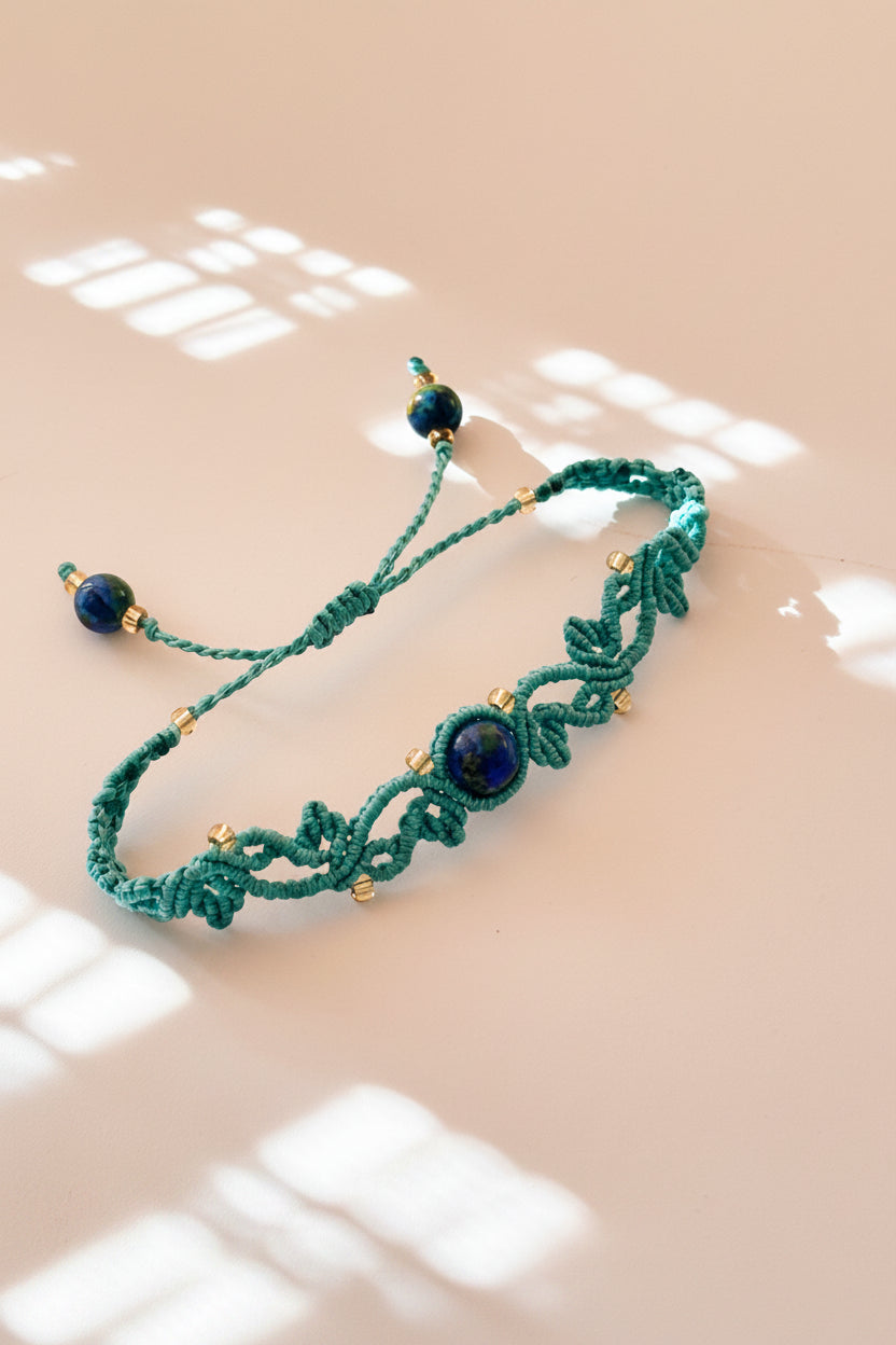 A handmade macrame bracelet in teal with dark blue beads and small gold-lined seed beads, displayed on a warm beige surface with soft window shadow patterns.