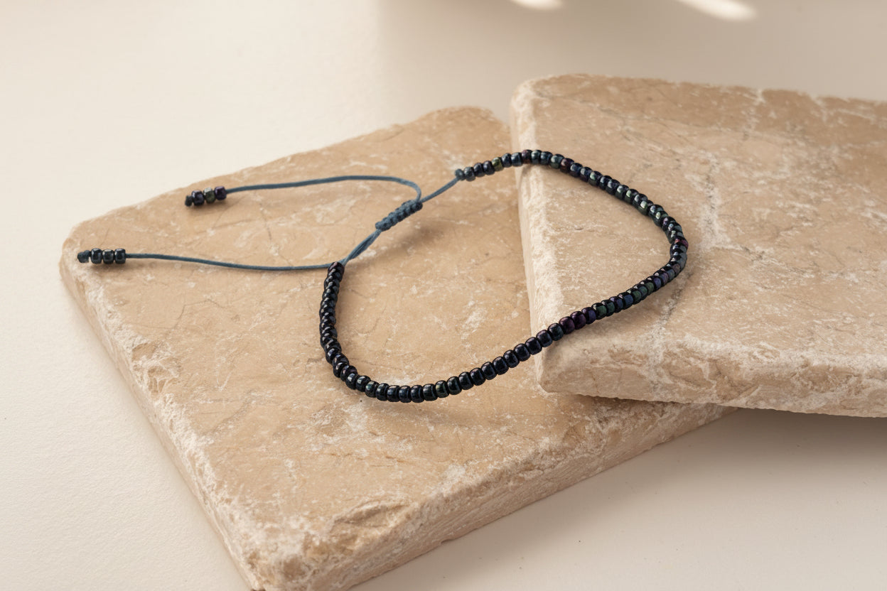 A handcrafted single strand bracelet with size 8/0 dark blue iridescent seed beads on a medium blue nylon cord with an adjustable slide closure, displayed on overlapping natural stone slabs against a white background