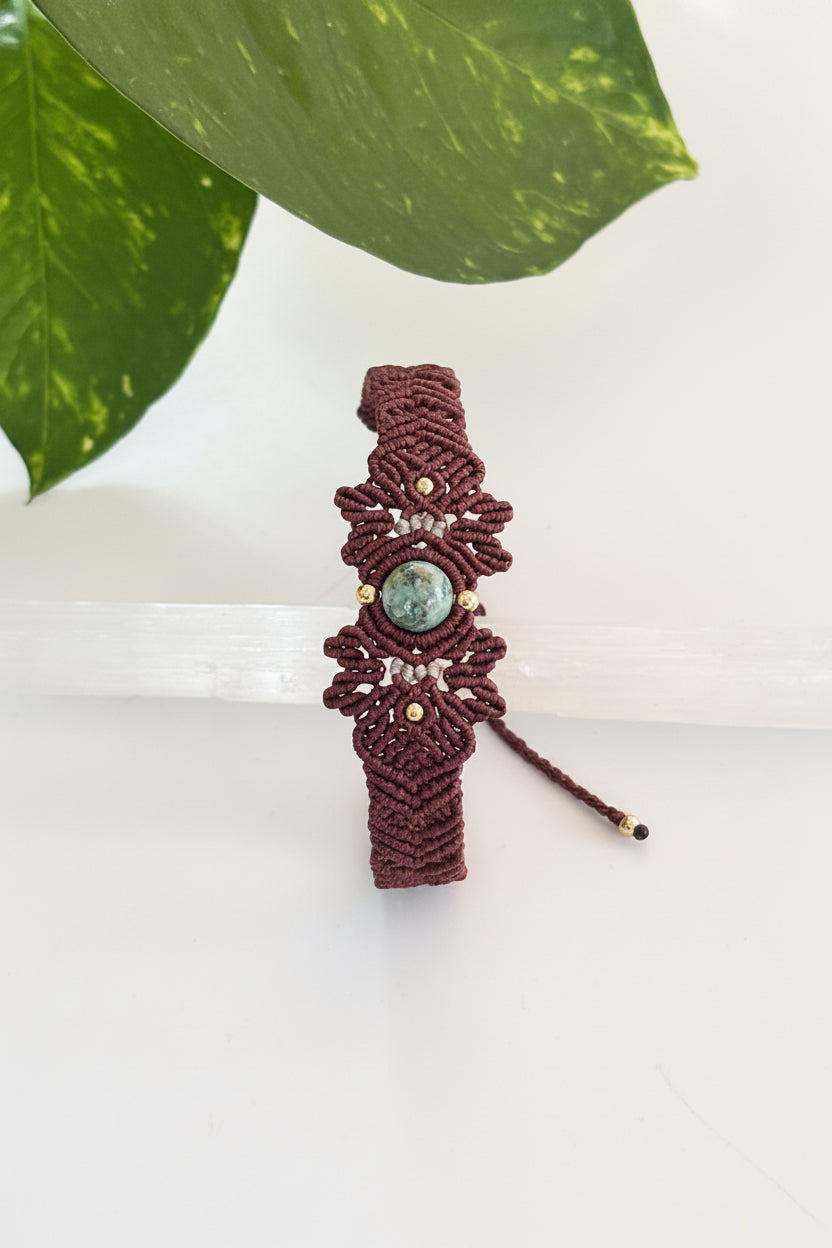 A handmade macrame bracelet in brown with a central round Indian Agate green stone and small brass beads, displayed on a clear glass cylinder with a green leaf in the background.