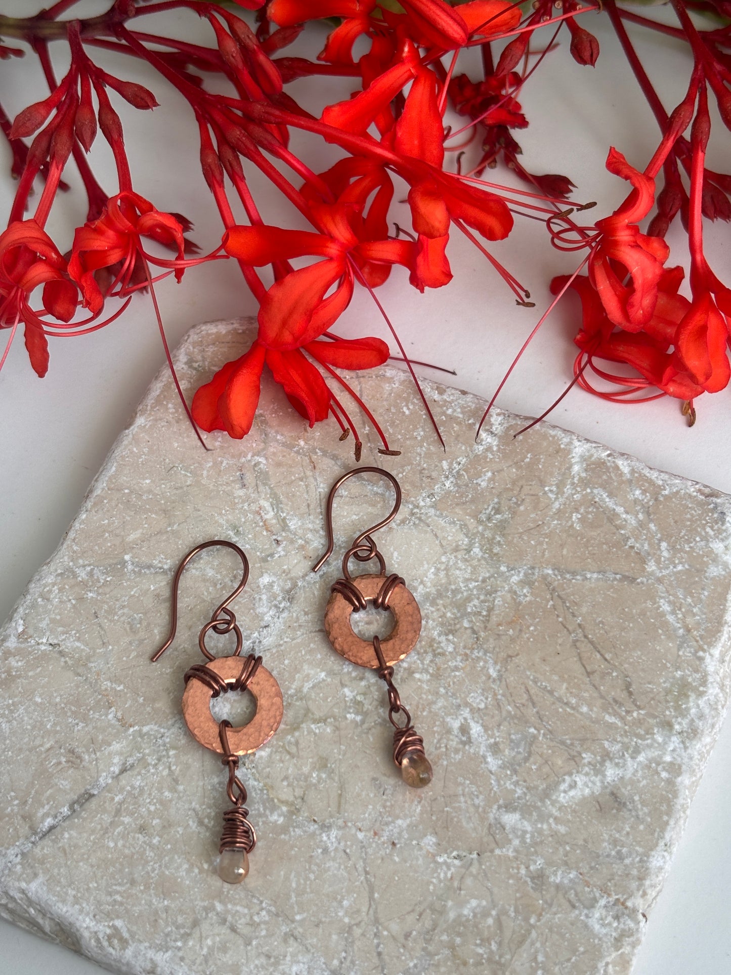 A pair of handmade copper earrings with circular ring charms and wrapped wire details, displayed on a textured stone slab with red flowers in the background.