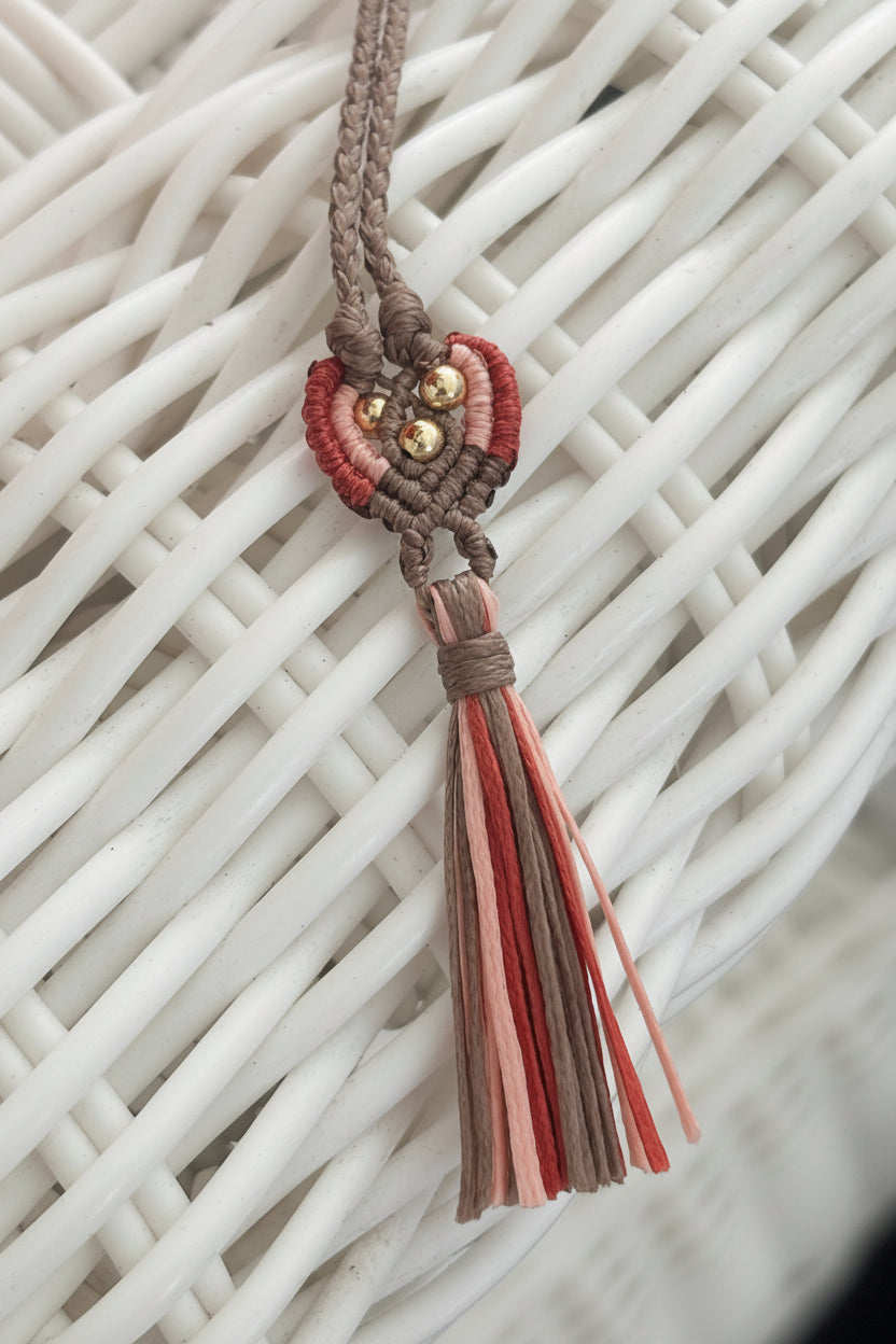 A handmade macrame tassel charm with a braided brown cord, rust, peach, and brown woven accents, brass beads, and a multi-colored tassel, resting on a white wicker surface.