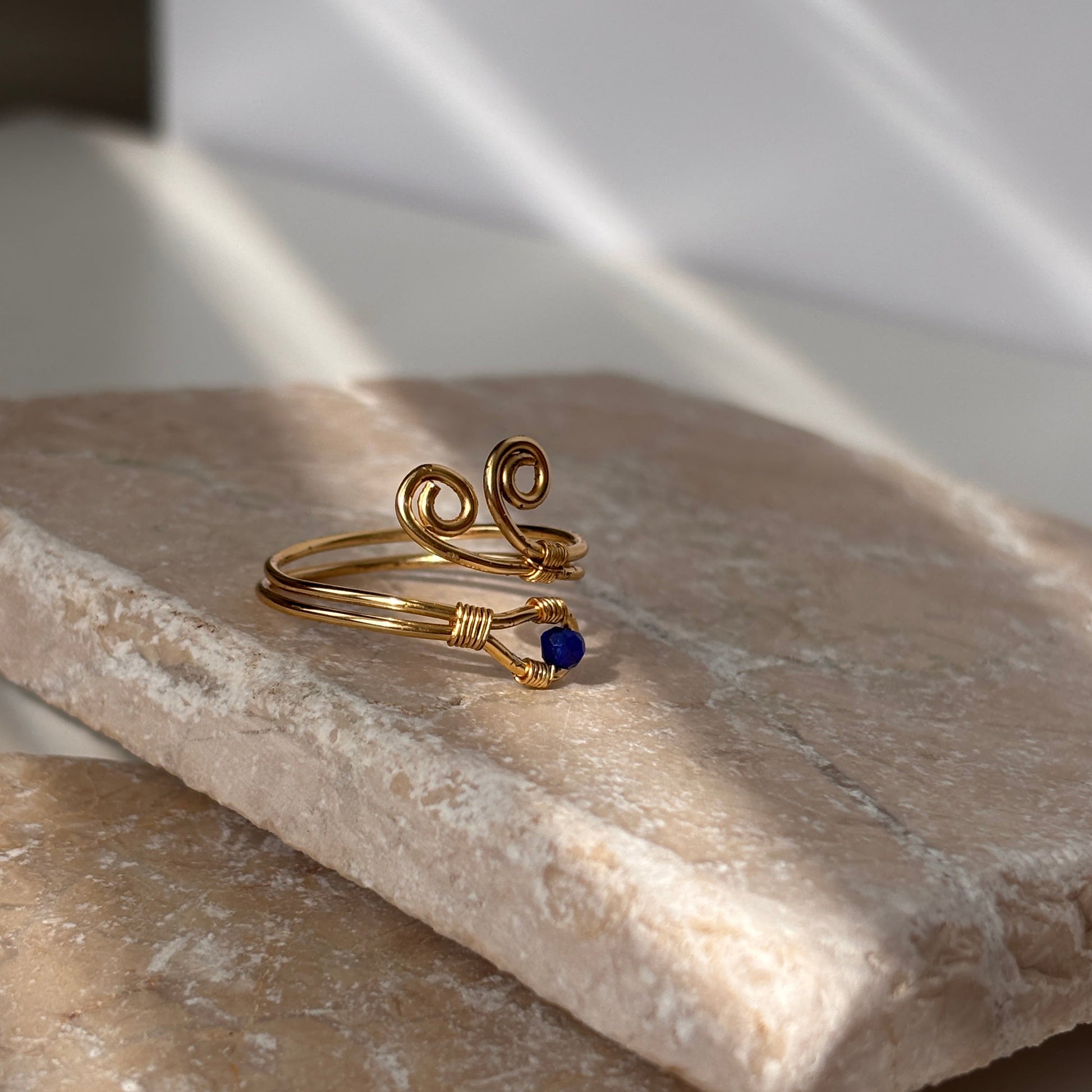A handcrafted gold-tone wire ring with a double spiral flourish and a 3mm deep blue faceted crystal bead wire-wrapped at the base, resting on a natural stone slab in soft natural sunlight against a white background