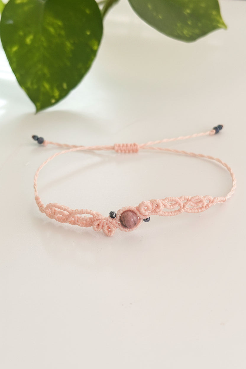 A light pink macrame bracelet with a central rhodonite stone bead and black iridescent crystal beads, resting on a light surface with a green leaf in the background.