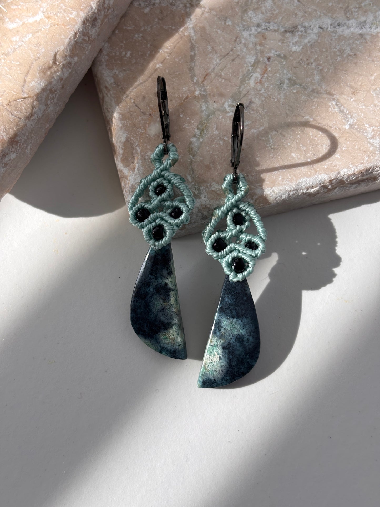 A pair of handcrafted macrame dangle earrings in teal nylon cord with intricate knotwork, black seed bead accents, and hand-selected Bacan stone teardrop pendants in deep teal and charcoal with natural mineral patterning, with gunmetal lever back ear wires, displayed on a white surface with a pink stone prop in natural sunlight