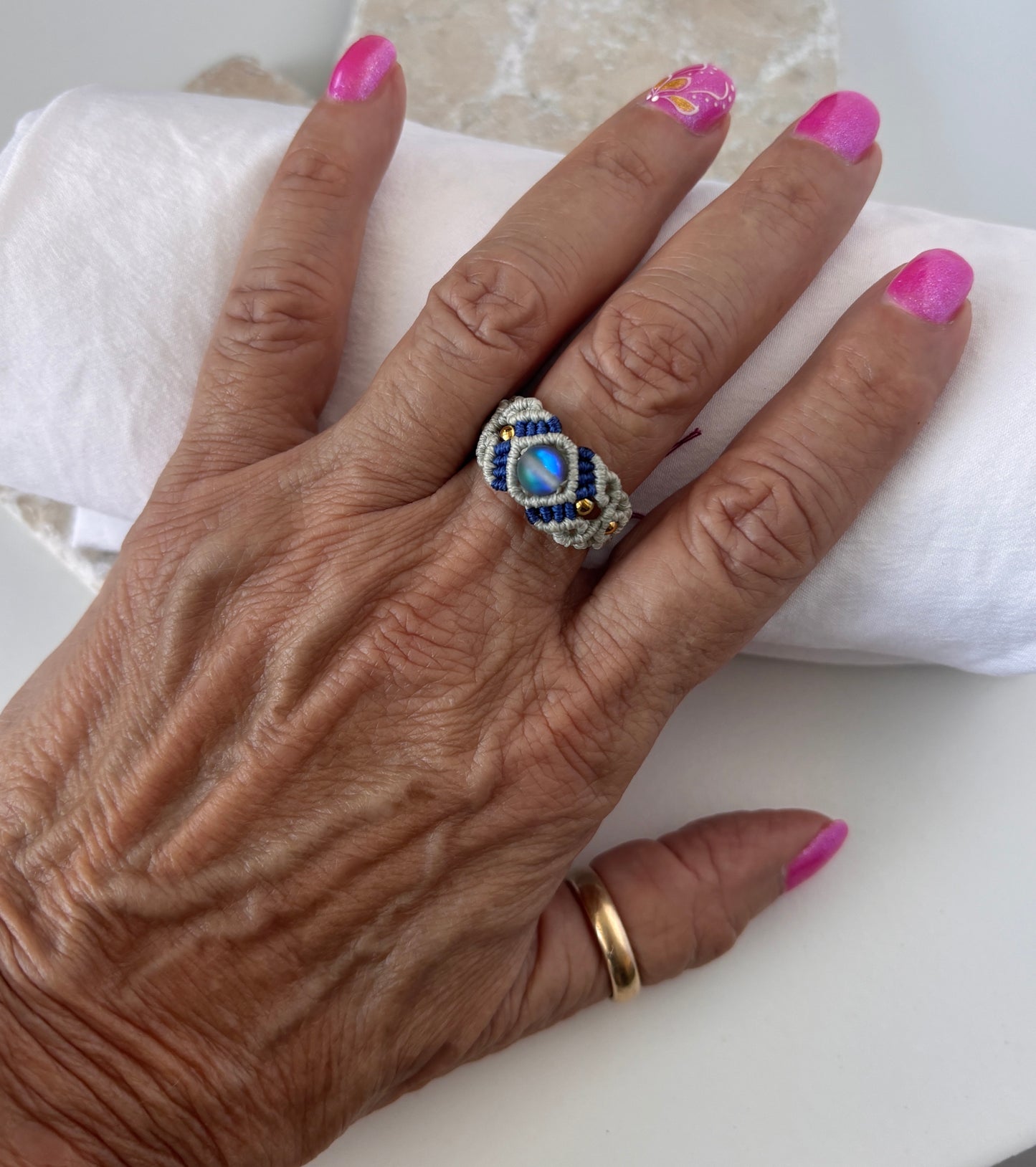 A handmade two-tone light gray and navy blue macrame cigar band ring with a shimmering iridescent mermaid glass bead and gold brass accent beads, worn on a finger with pink nails resting on a white surface.