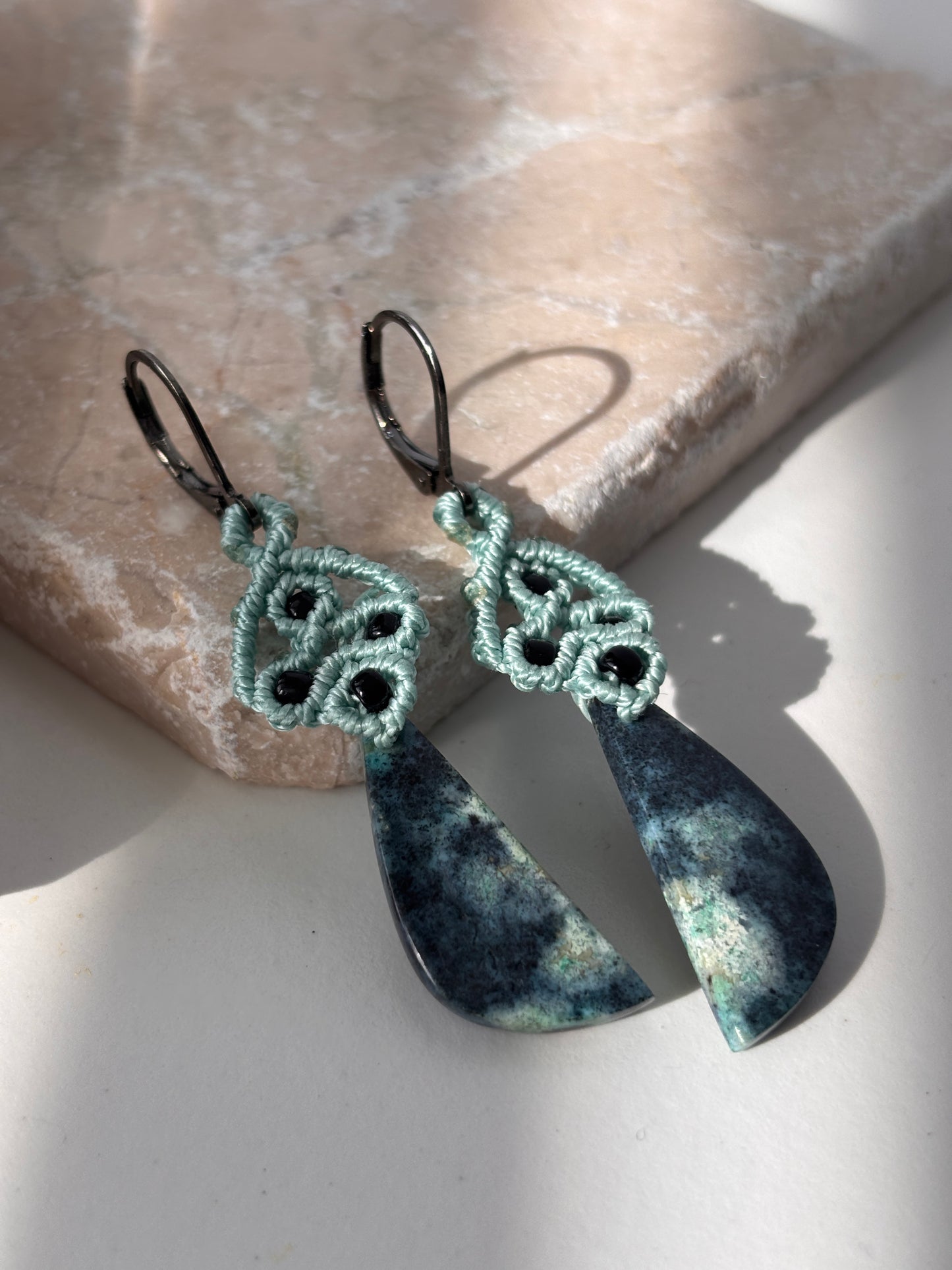 A pair of handcrafted macrame dangle earrings in teal nylon cord with intricate knotwork, black seed bead accents, and hand-selected Bacan stone teardrop pendants in deep teal and charcoal with natural mineral patterning, with gunmetal lever back ear wires, leaning against a pink stone surface in natural sunlight.