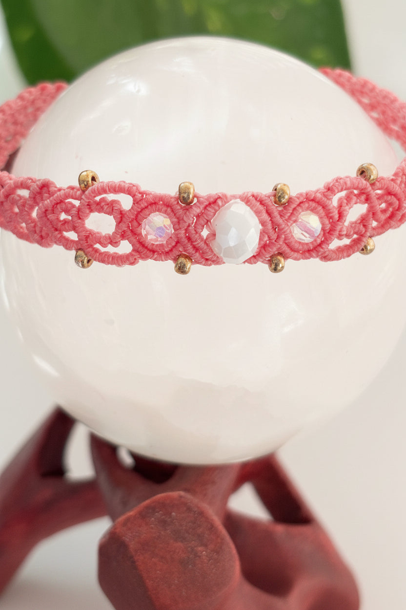 A salmon pink macrame bracelet with brass beads and a central faceted iridescent crystal framed on each side by small iridescent pink round faceted beads, displayed wrapped around a large white crystal sphere on a wooden stand.